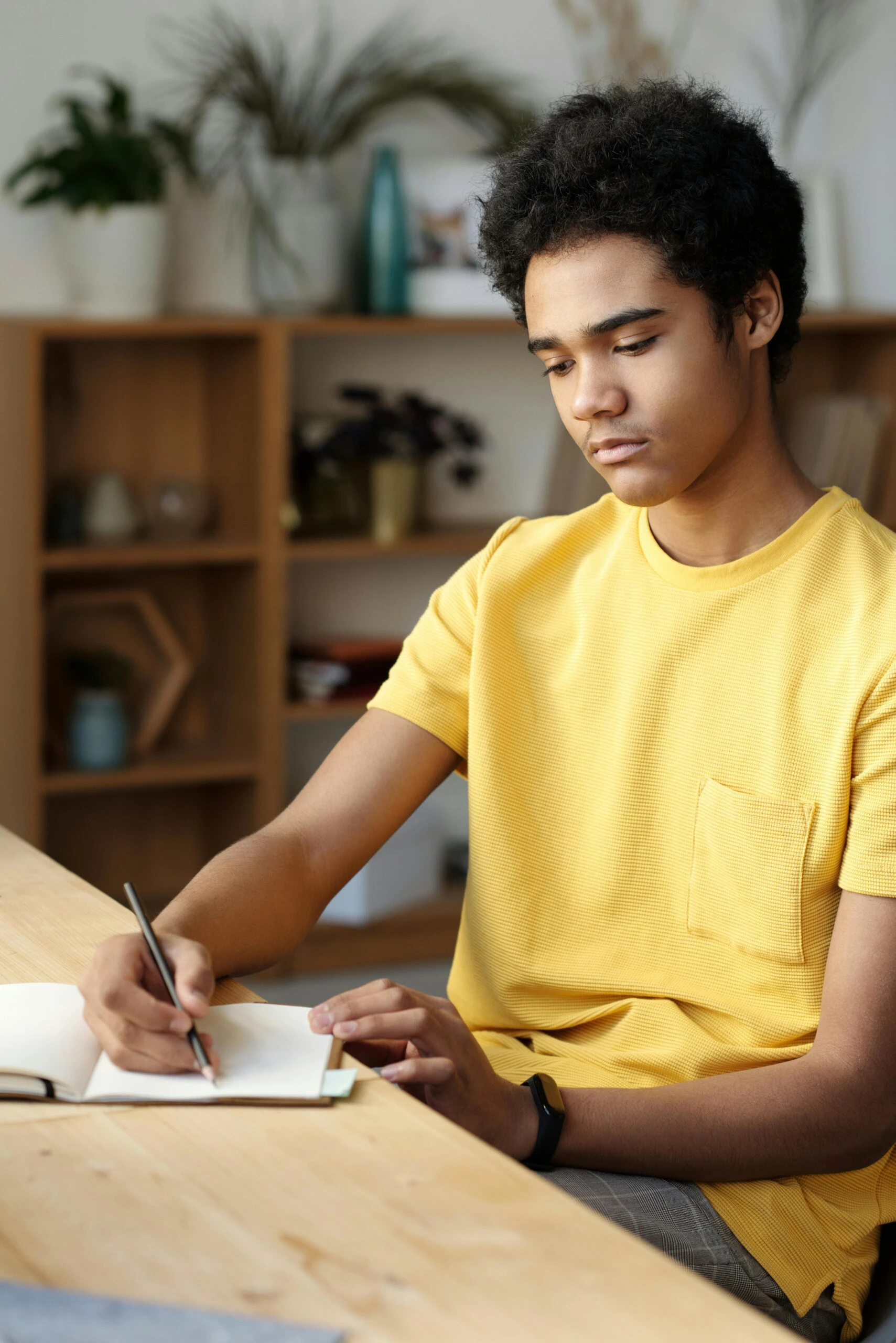 Focused teen boy studying at home in a yellow shirt, writing notes in a bright room. Education concept.